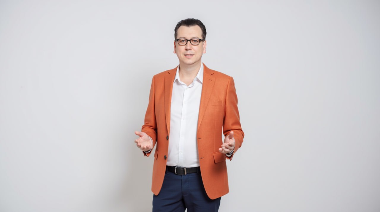 Артем Чусов - генеральный директор в оранжевом пиджаке улыбается и приветственно разводит руки.