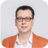 Артем Чусов - генеральный директор в оранжевом пиджаке улыбается и приветственно разводит руки.